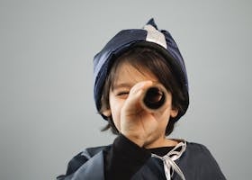 A child in a wizard costume looks through a telescope against a gray background.